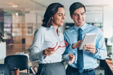 Frau und Mann in Business-Kleidung stehen nebeneinander in einem Büro und schauen sich etwas auf einem Tablet an. Beide lächeln. Hinter Ihnen sind weitere Schreibtische und eine große Glasfront.